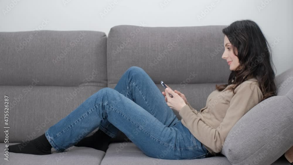 Woman browsing internet on mobile phone while lying on comfortable couch in living room at home. Stock video clip of beautiful middle aged female model using modern smartphone to read news Woman browsing internet on mobile phone while lying on comfortable couch in living room at home. Stock video clip of beautiful middle aged female model using modern smartphone to read news