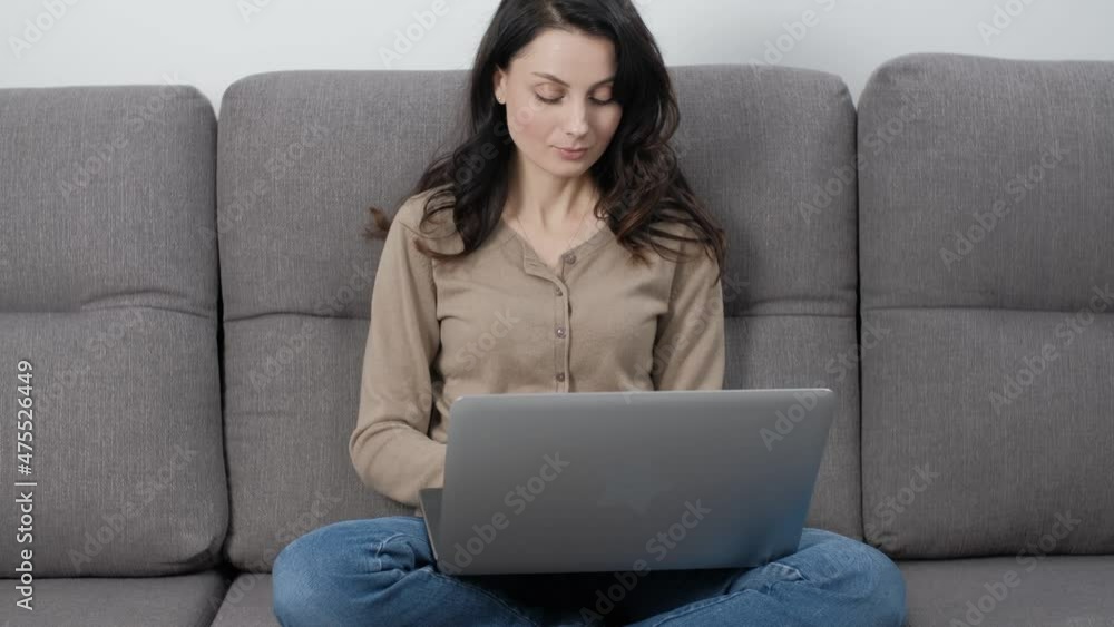 Stock video of freelancer woman working from home with laptop computer. Beautiful young adult model with long dark hair typing text on notebook keyboard while sitting on comfortable sofa in room Stock video of freelancer woman working from home with laptop computer. Beautiful young adult model with long dark hair typing text on notebook keyboard while sitting on comfortable sofa in room