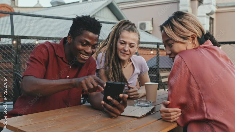 Medium shot of happy young diverse friends laughing while looking at smartphone spending summertime together at outdoor rooftop cafe Medium shot of happy young diverse friends laughing while looking at smartphone spending summertime together at outdoor rooftop cafe