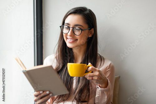 Carefree latin lady with cup of tea reading favorite book while resting in cafe near window  empty space