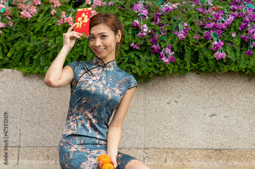 Single brown hair slim Asian woman dressed in festive Cheongsam for Lunar Chinese New Year celebration. Female holding mandarin oranges red pack. Translation : Wish you all the wealth and prosperity.