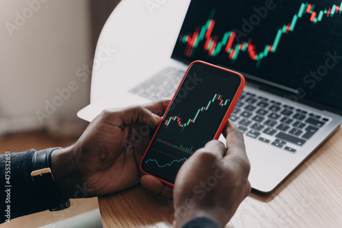 Male hands holding smartphone next to computer screen analyzing financial stock market charts