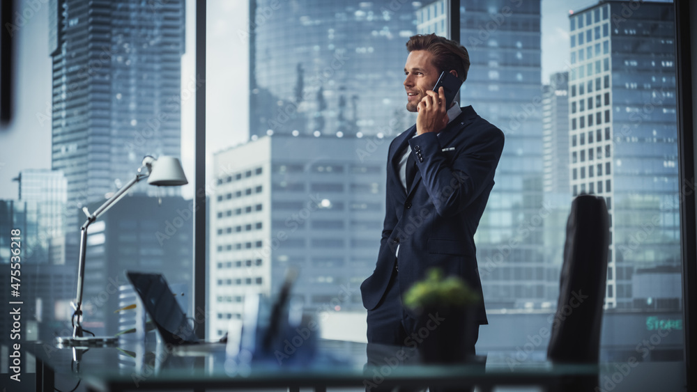 Fototapeta premium Confident Young Businessman in a Suit Standing in Modern Office, Talking on a Phone, Looking out of the Window on Big City with Skyscrapers. Successful Finance Manager Planning Work Projects.