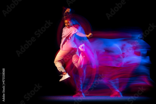Emotional young girl in white costume dancing hip-hop dance isolated on dark background at dance hall in neon mixed light.