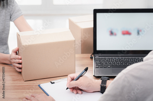 Post man use a pen write on file paper document and check online system to send parcels and women hold paper box laptop on table : delivery concept work