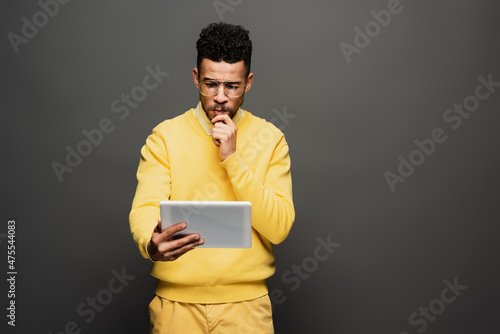 pensive african american man in glasses using digital tablet on dark grey.