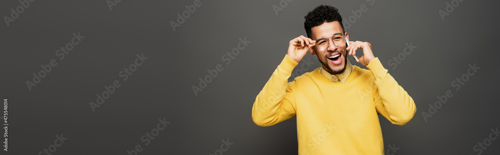 happy african american man adjusting glasses and talking on smartphone on dark grey, banner. happy african american man adjusting glasses and talking on smartphone on dark grey, banner.