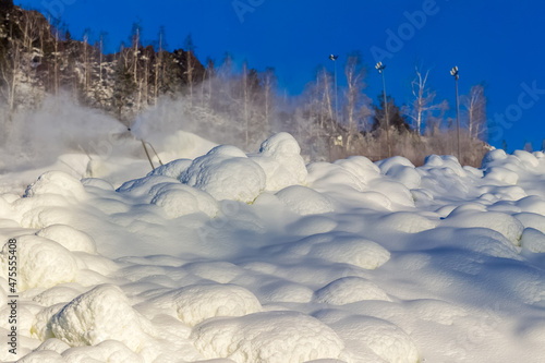 Snowdrifts and flying snow from a snow cannon on the mountainside against the background of trees and blue sky in winter