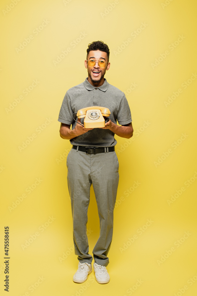 full length of excited african american man in sunglasses and grey tennis shirt holding retro telephone on yellow. full length of excited african american man in sunglasses and grey tennis shirt holding retro telephone on yellow.