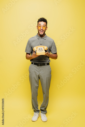 full length of excited african american man in sunglasses and grey tennis shirt holding retro telephone on yellow. © LIGHTFIELD STUDIOS