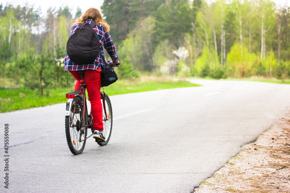 Kiev, Ukraine. May 1, 2020. A girl, young woman in checkered shirt, red pants, black backpack riding bicycle on a country paved road in woods, park in spring, summer. Journey alone, healthy lifestyle.