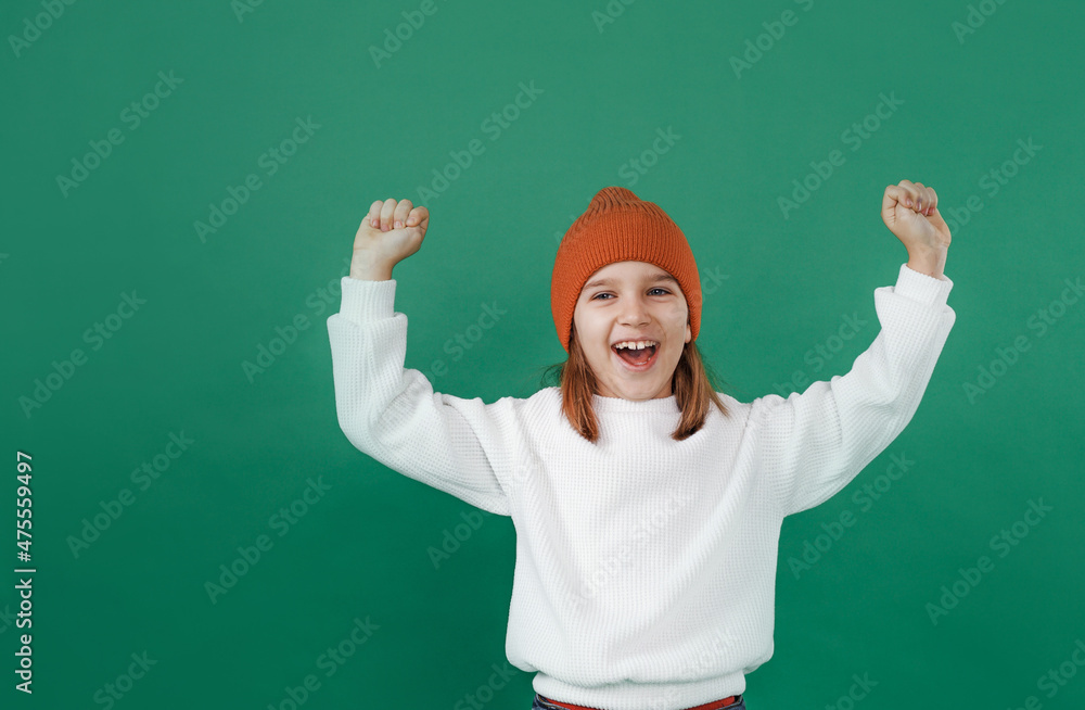 A preschool child in a white sweater and hat smiles, raises his hands up and laughs on an isolated green background.