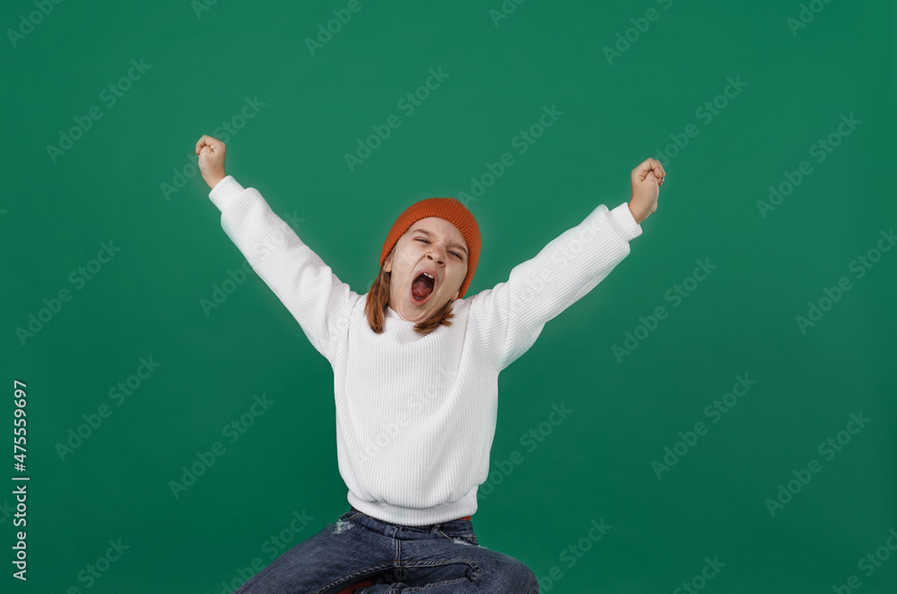 a young boy in a white sweater and hat yawns, opening his mouth wide and raising his hands up against a green background.