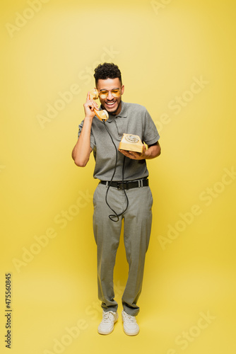 full length of happy african american man in sunglasses and grey tennis shirt talking on retro telephone on yellow.