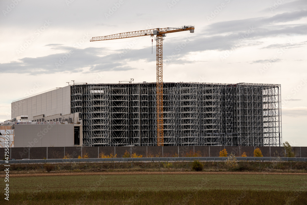Warehouse under construction without external wall. Metal shelves of a future logistics center. Construction of a steel building, in the foreground a yellow construction crane Warehouse under construction without external wall. Metal shelves of a future logistics center. Construction of a steel building, in the foreground a yellow construction crane
