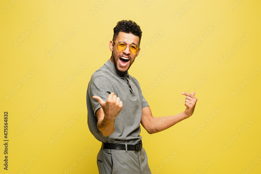 joyful african american man in sunglasses and grey tennis shirt gesturing isolated on yellow. joyful african american man in sunglasses and grey tennis shirt gesturing isolated on yellow.