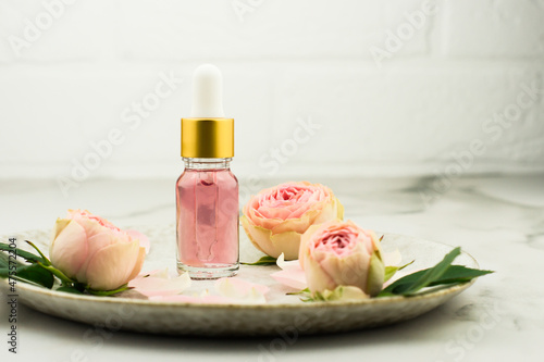 a glass bottle with a dropper pipette with essential oil of rose petals on a marble table opposite a white brick wall. rose buds on a ceramic plate.