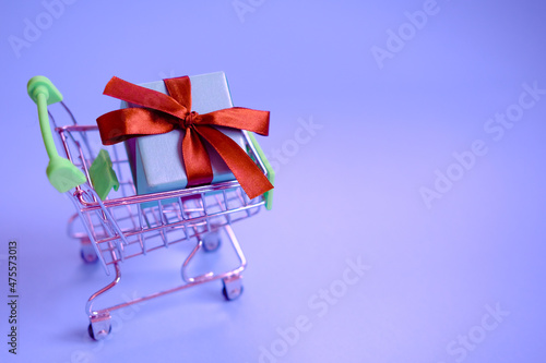 A present box with a ribbon in a shopping trolley cart on very pari background . Concept - buying a gift ,online and sale. Christmas, valentines ,women day .Copy space, mockup.