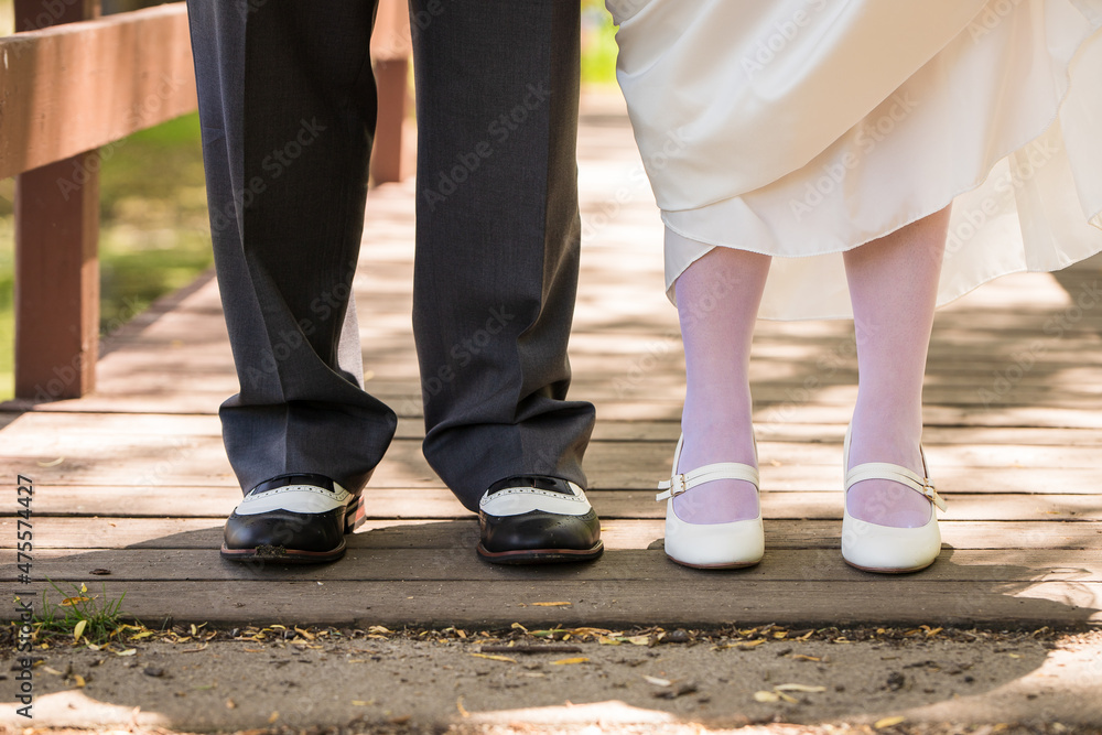 The feet of a bride and groom standing together on a bridge on their wedding day. The bride lifts her dress to reveal her tights and white shoes.