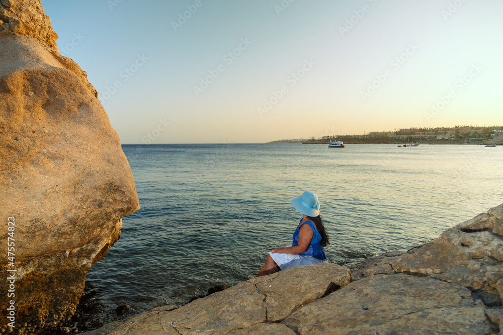 A young woman in a white skirt and a sun hat on the seashore sits on a stone admiring nature with her back to the camera.