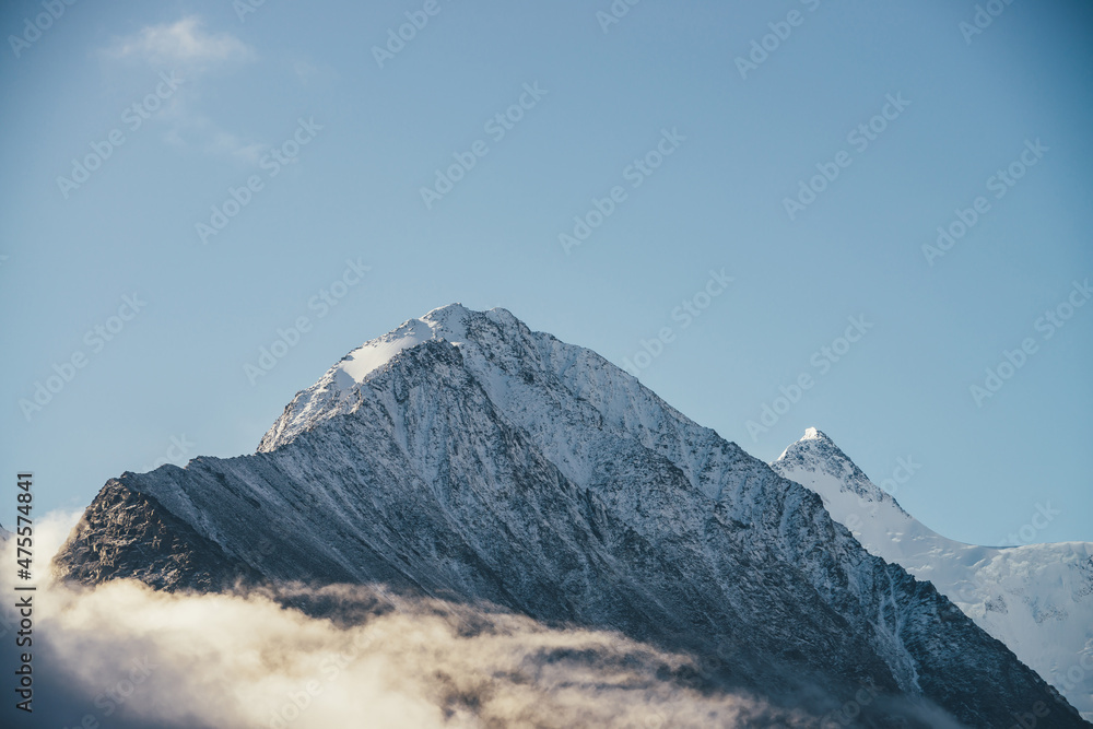 Beautiful view of snow-capped mountains above thick clouds in sunshine. Scenic bright mountain landscape with white-snow peak among dense low clouds in blue sky. Wonderful scenery with snowy pinnacle.