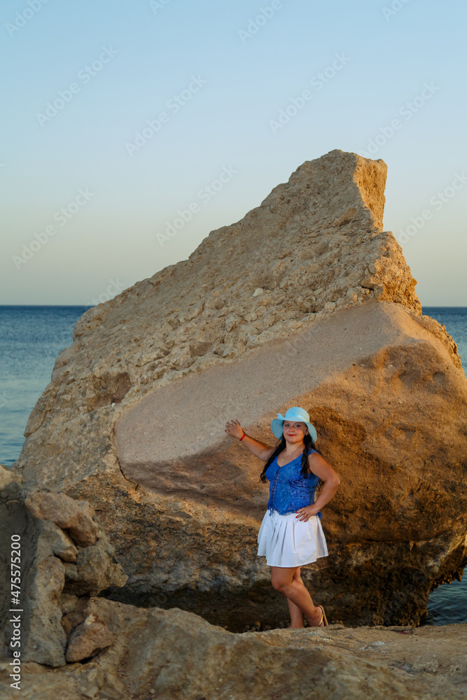 Naklejka premium A young woman in a white skirt and a sun hat stands on the shore among the rocks against the backdrop of the sea at sunset.
