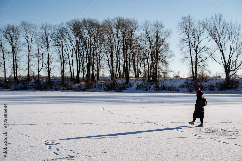 A young girl in a bright hat walks on a frozen lake. Delight and joy from the winter landscape with bright sun, clear sky. Walk in nature.