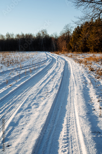 Winter landscape. A winding deserted road running forward through a field. Snow-covered forest in the sunset rays of the winter sun. Car tracks in the snow