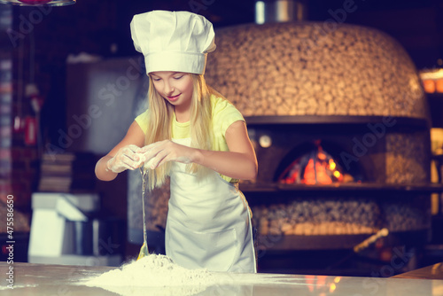A baker girl in a large uniform hat adds an egg to the flour.