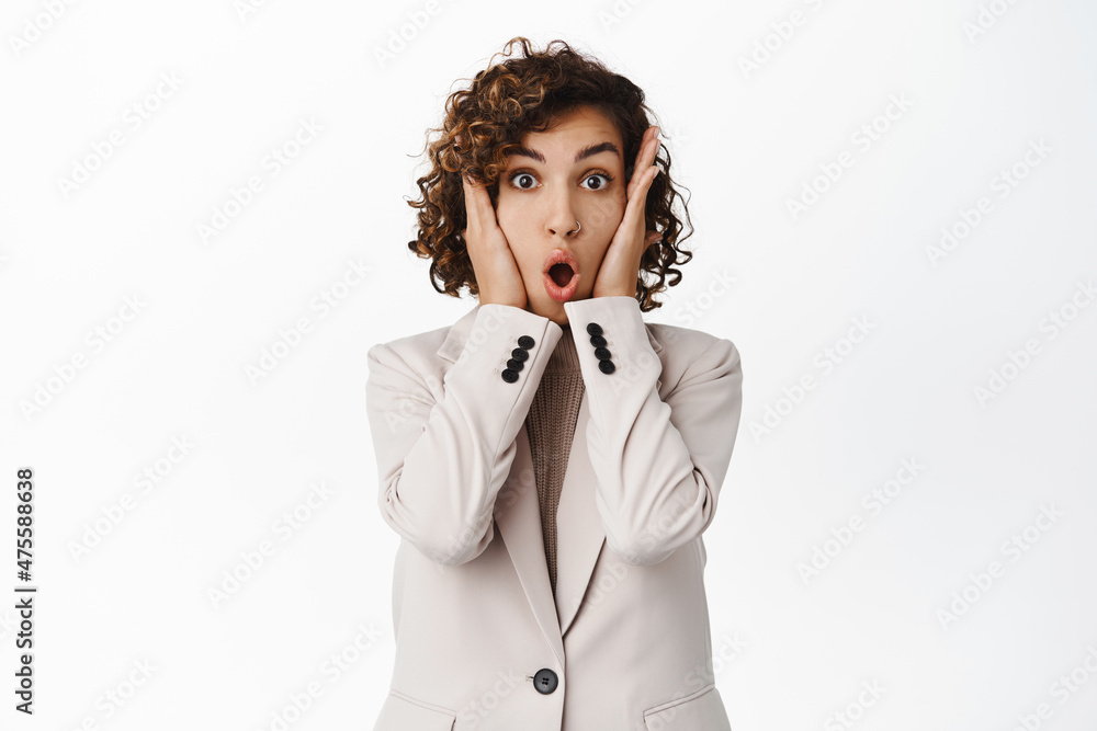 Shocked and amazed young corporate woman looking speechless, gasping and saying wow, standing over white background