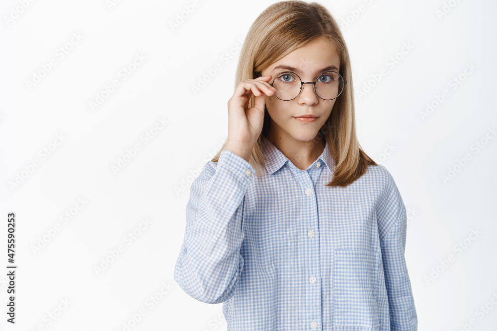 Cute clever blond girl in glasses looking smart at camera, standing in shirt over white background