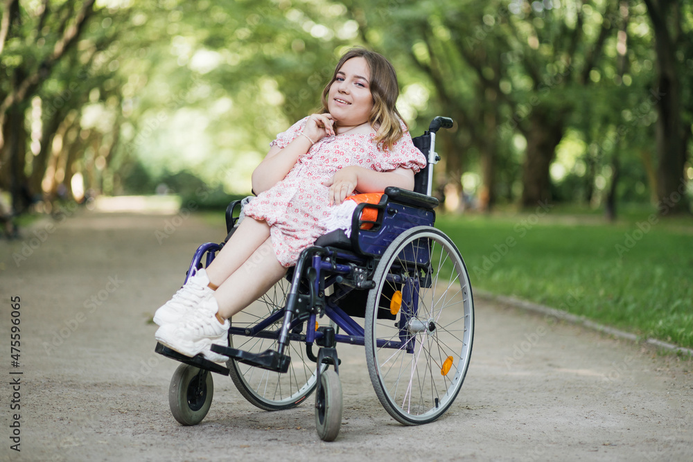 Portrait of pleasant young woman with spinal muscular atrophy smiling on camera among green summer park. Female person who using wheelchair. Concept of people with disability. Portrait of pleasant young woman with spinal muscular atrophy smiling on camera among green summer park. Female person who using wheelchair. Concept of people with disability.