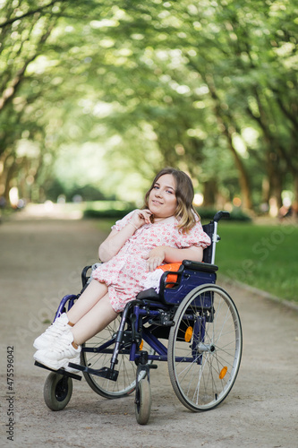Portrait of pleasant young woman with spinal muscular atrophy smiling on camera among green summer park. Female person who using wheelchair. Concept of people with disability. © sofiko14