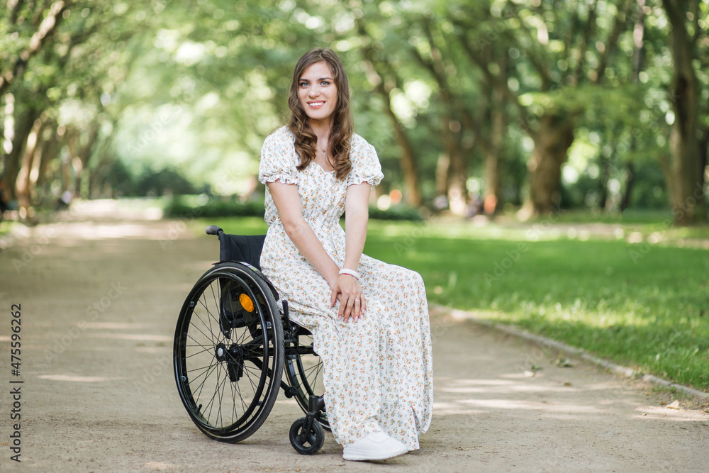 Smiling woman who lives with disability posing on camera among green summer park. Caucasian brunette wearing stylish dress with floral print. Positive emotions of young lady who using wheelchair.