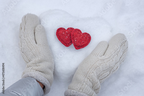 Women's hands in white knitted mittens with a red heart made of snow on a winter day. Love concept. Valentine day background.