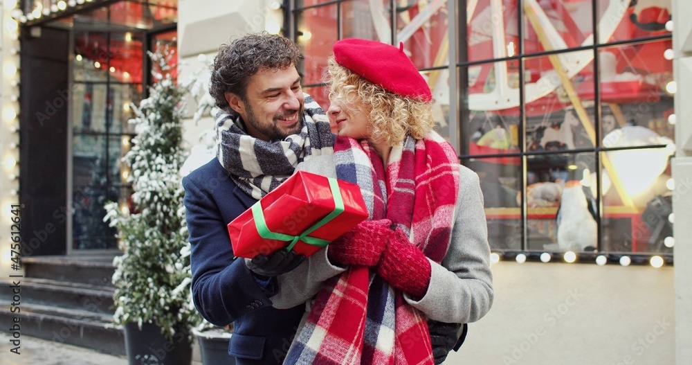 Obraz premium Man smiling and hugging his girlfriend and gives Christmas gift on decorated street. Christmas mood concept. Unexpected surprise.