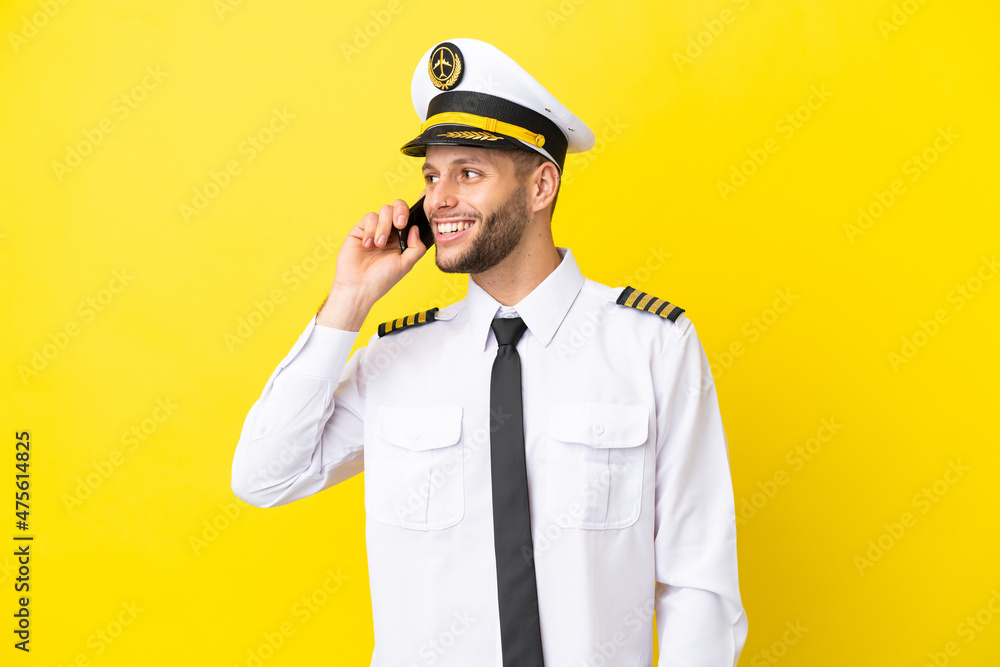 Airplane caucasian pilot isolated on yellow background keeping a conversation with the mobile phone Airplane caucasian pilot isolated on yellow background keeping a conversation with the mobile phone