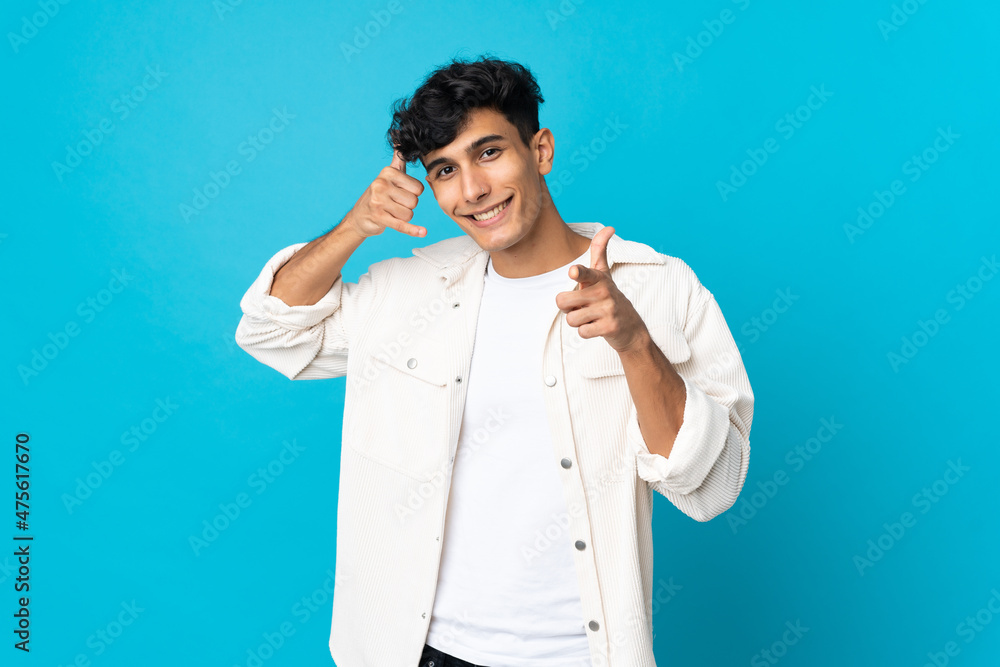 Young Argentinian man isolated on background making phone gesture and pointing front Young Argentinian man isolated on background making phone gesture and pointing front
