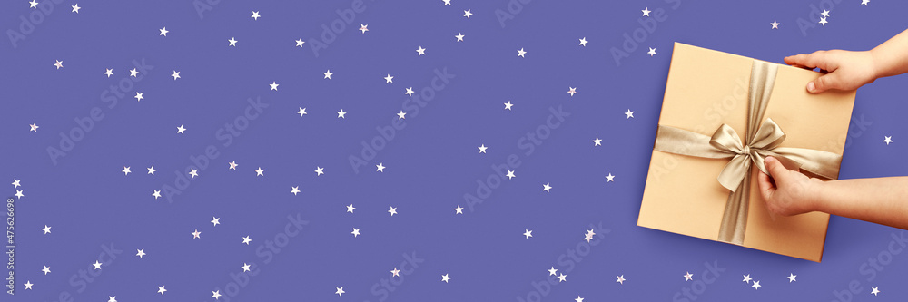 Banner made with baby hands are holding a box with a gift. Cute festive very peri color background with sparkles. Conceptual backdrop for christmas, new year or birthday. Banner made with baby hands are holding a box with a gift. Cute festive very peri color background with sparkles. Conceptual backdrop for christmas, new year or birthday.