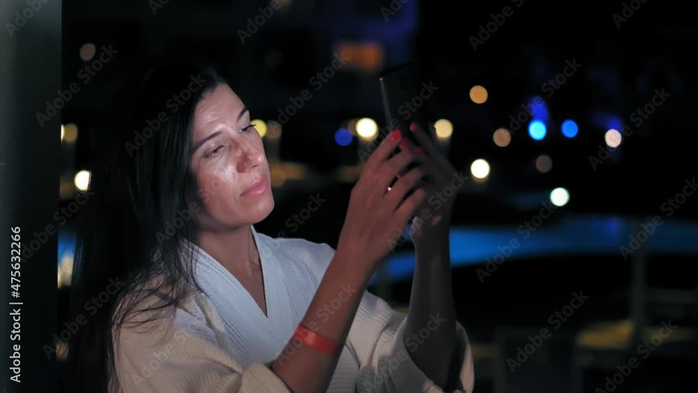 before bedtime. woman in white bathrobe, uses smartphone, on an open balcony, late in the evening or at night. her face is illuminated by smartphone screen light. blurry city lights can be seen in before bedtime. woman in white bathrobe, uses smartphone, on an open balcony, late in the evening or at night. her face is illuminated by smartphone screen light. blurry city lights can be seen in