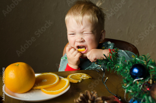 A little boy eats and winces an orange at a New Year's table in pajamas home. Child is waiting for the holidays and Christmas concept