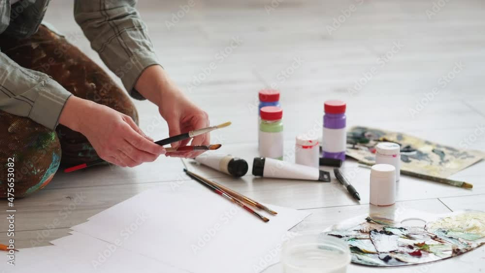Painting equipment. Female artist. Artistic tools. Unrecognizable woman choosing brushes with paint bottles sitting wooden floor in light room interior. Painting equipment. Female artist. Artistic tools. Unrecognizable woman choosing brushes with paint bottles sitting wooden floor in light room interior.