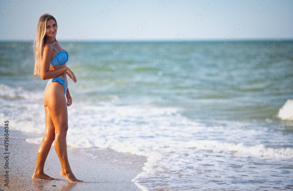 A girl with blond hair in a bluish swimsuit walks along the beach