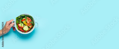 A female hand holds a bowl of vegetable salad on a blue background. Top view, flat lay. Banner
