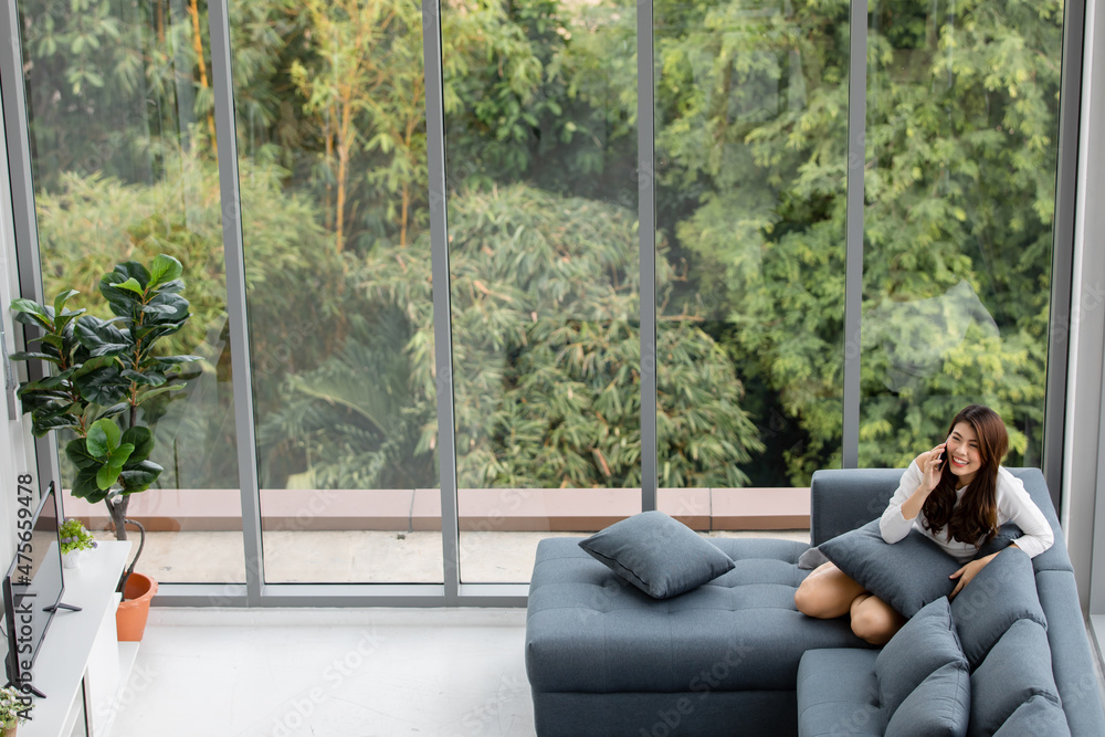 Asian female housewife in casual outfit sitting on cozy sofa couch with pillows resting relaxing smiling talking conversation on call with friend in comfortable living room with garden view at home Asian female housewife in casual outfit sitting on cozy sofa couch with pillows resting relaxing smiling talking conversation on call with friend in comfortable living room with garden view at home