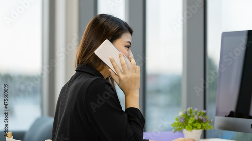 Asian happy successful female businesswoman in black suit sitting on chair typing keyboard computer at working desk in room with cozy sofa couch and bookshelf in beautiful garden view home office