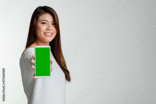 Portrait studio shot Asian young beautiful female model in long sleeve shirt show naked shoulder standing smiling holding presenting black blank screen smartphone look at camera on white background