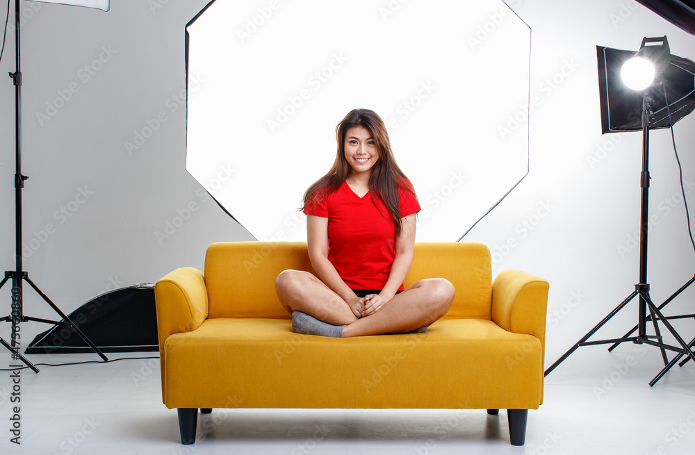 Portrait studio shot of Asian pretty thoughtful female model in red shirt sitting thinking on cozy yellow sofa couch on photographing shooting backstage set with softbox lighting reflector spotlight Portrait studio shot of Asian pretty thoughtful female model in red shirt sitting thinking on cozy yellow sofa couch on photographing shooting backstage set with softbox lighting reflector spotlight