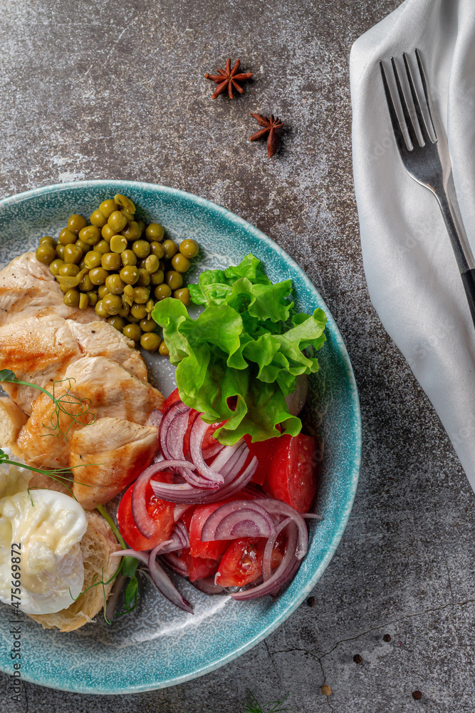 Serving a dish from the restaurant menu. Chicken breast with vegetables and greens, tomatoes, onions, omelettes and green peas on a plate against the background of a gray stone table