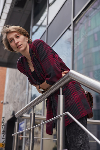young woman with short hair in red checkered jacket outdoors in front of office building. portrait of caucasian female leaning on railings at street